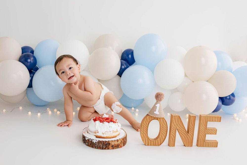 Baby boy crawling near a smash cake and ONE sign surrounded by blue and white balloons for a first birthday.