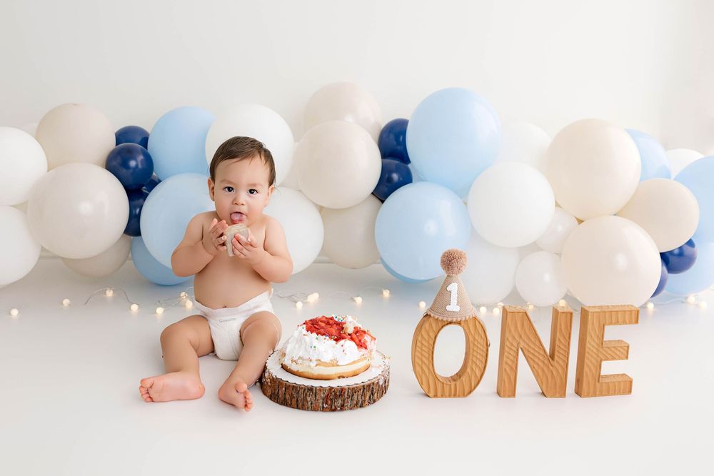 Baby boy celebrates first birthday smash cake with balloons and ONE sign.
