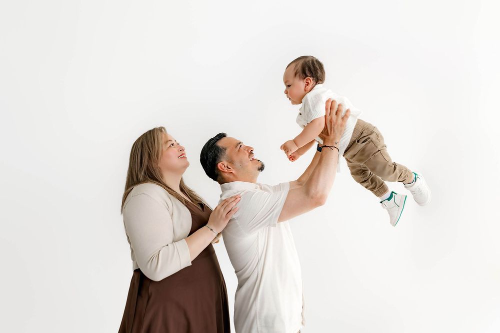 A joyful family with a mother, father, and baby, as dad lifts the smiling infant in a bright white studio.