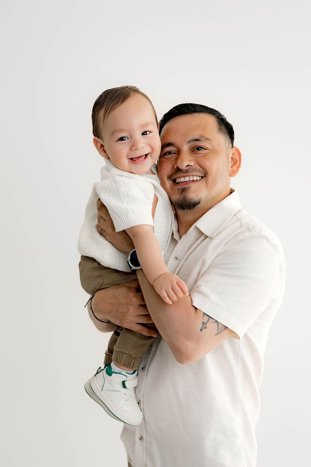 A smiling man with a tattoo holds a laughing baby dressed in white and olive, posing together against a white background.