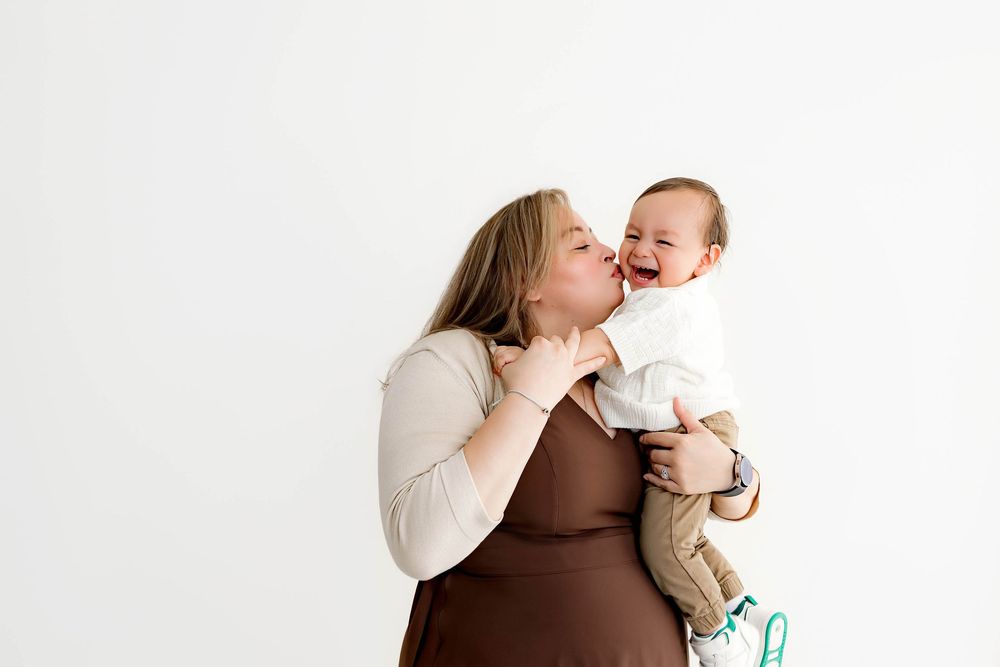 A woman in a brown dress holds a laughing baby while giving a gentle kiss on its cheek against a white background.