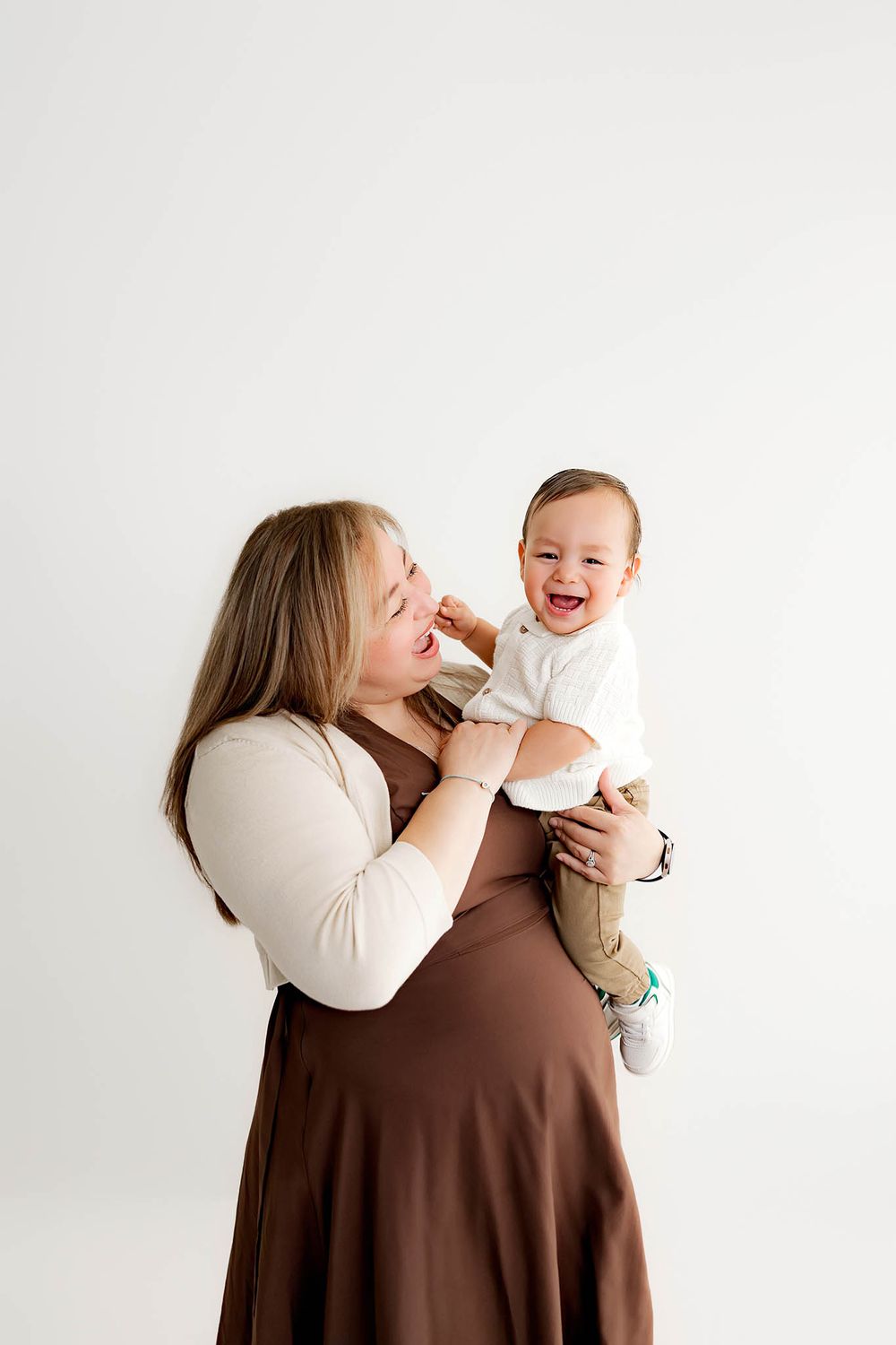 A pregnant woman in a brown dress holds a laughing baby boy in a bright white studio setting.