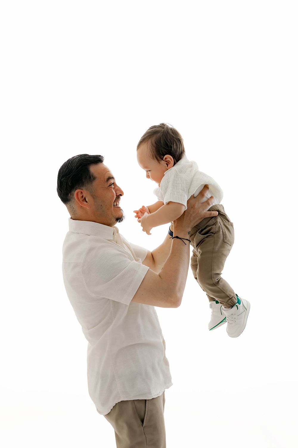 A joyful father lifts his smiling baby boy in the air against a white background.