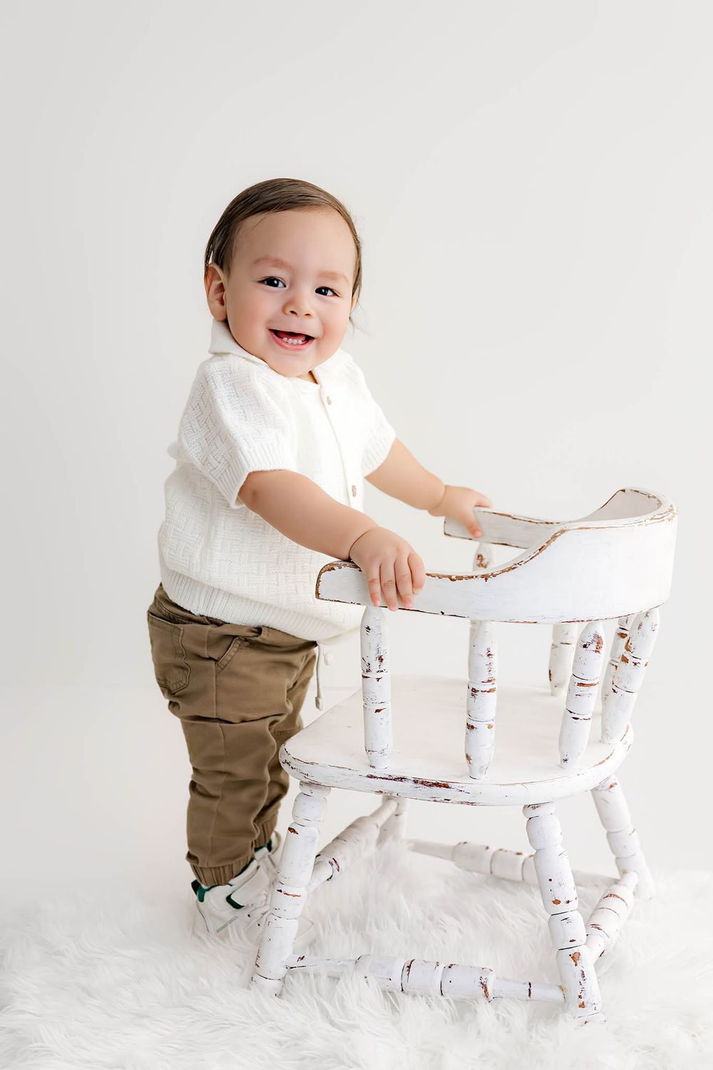 Cheerful toddler in white shirt and khaki pants standing by a rustic white chair on fluffy rug.
