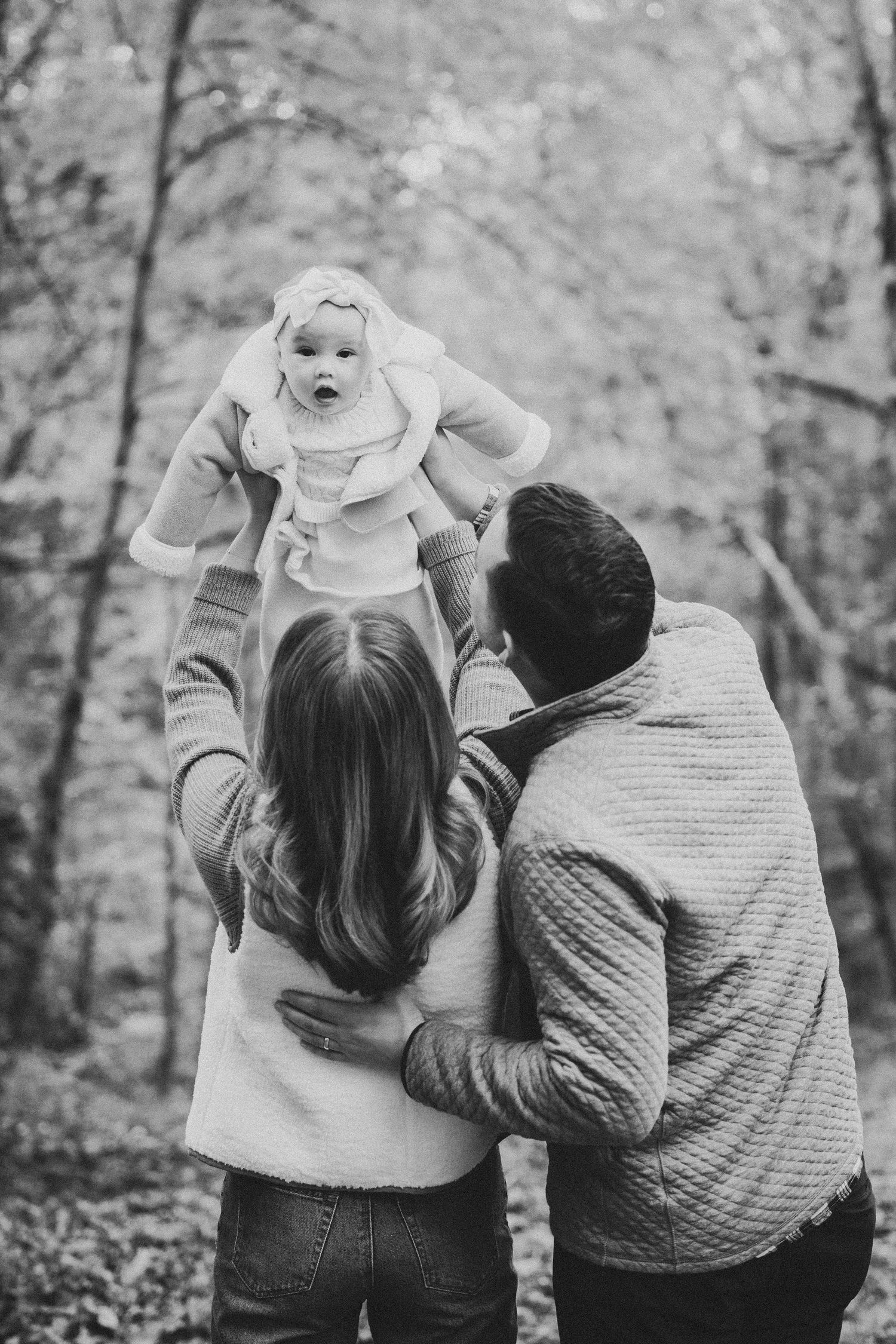 Black and white photo of parents lifting their baby in an autumn forest, sharing a tender family moment outdoors.