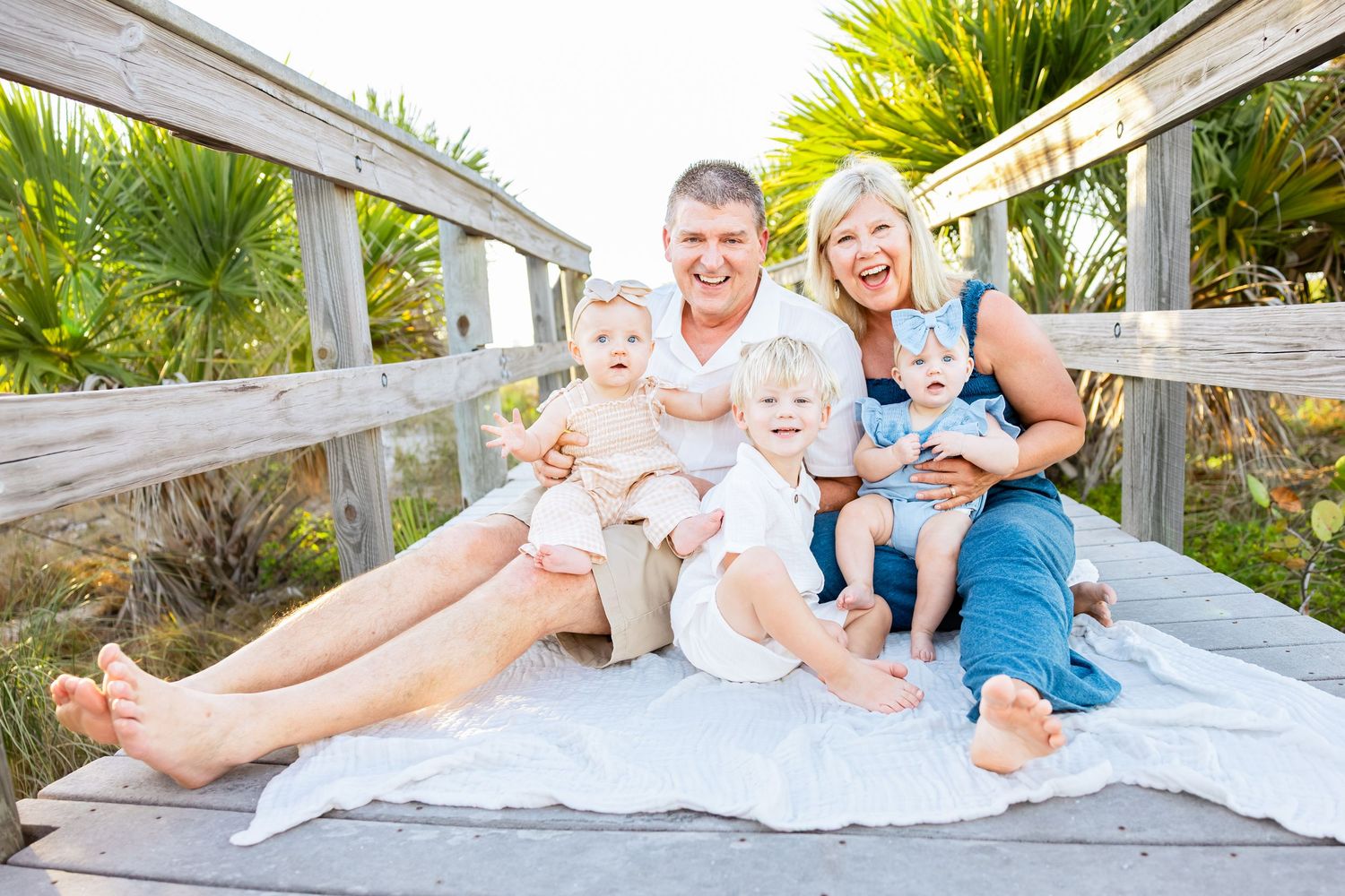 Happy family with three young children smiling on a wooden boardwalk near a tropical beach on a sunny day.
