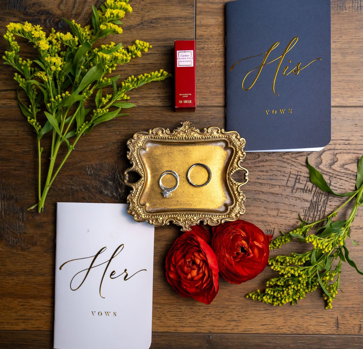 Bride's vow books, wedding rings on a gold tray, red lipstick, red ranunculus and yellow wildflowers on a rustic wooden table.