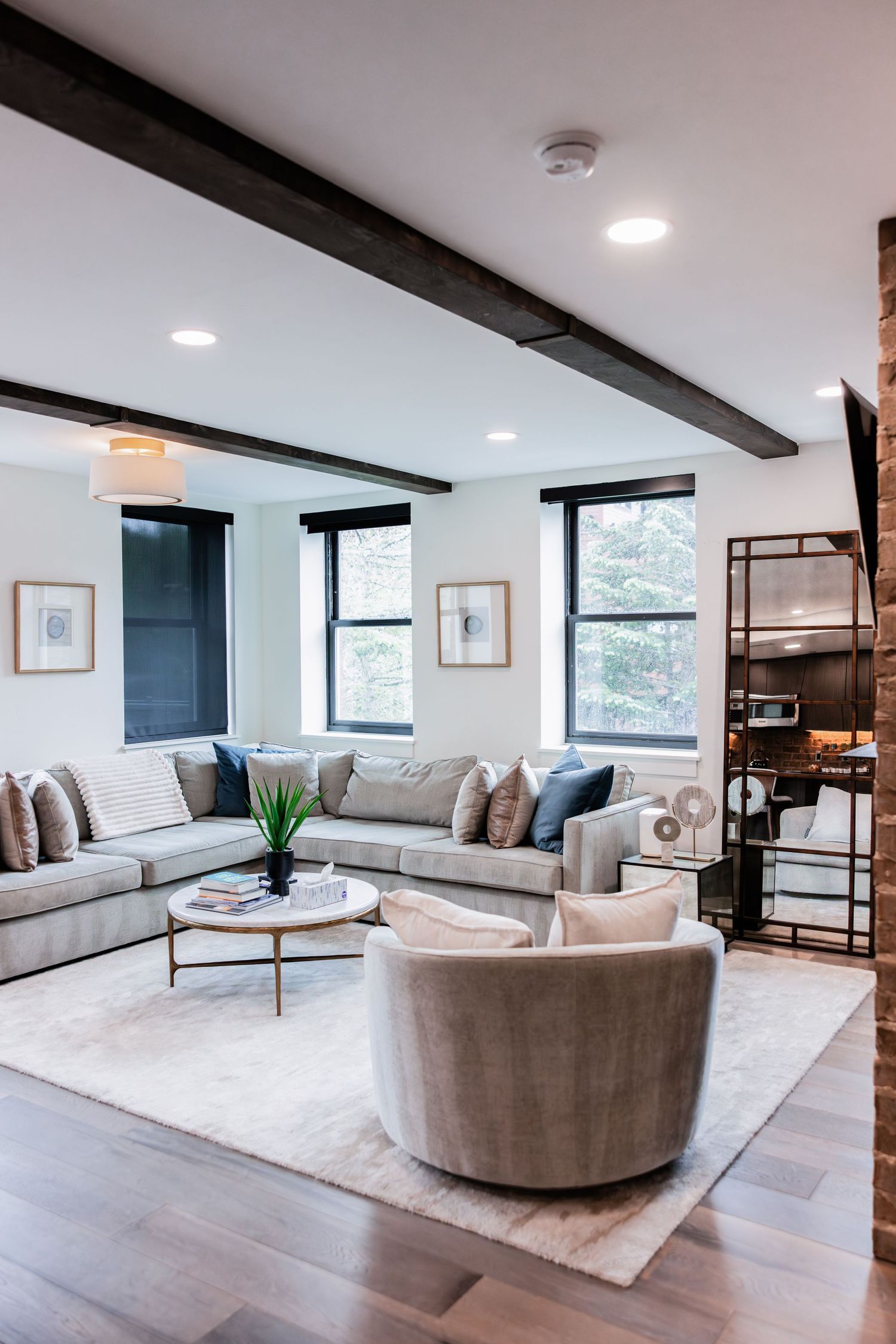 Modern living room with a gray sectional sofa, swivel armchair, round coffee table, exposed wood beams, and large black-framed windows.