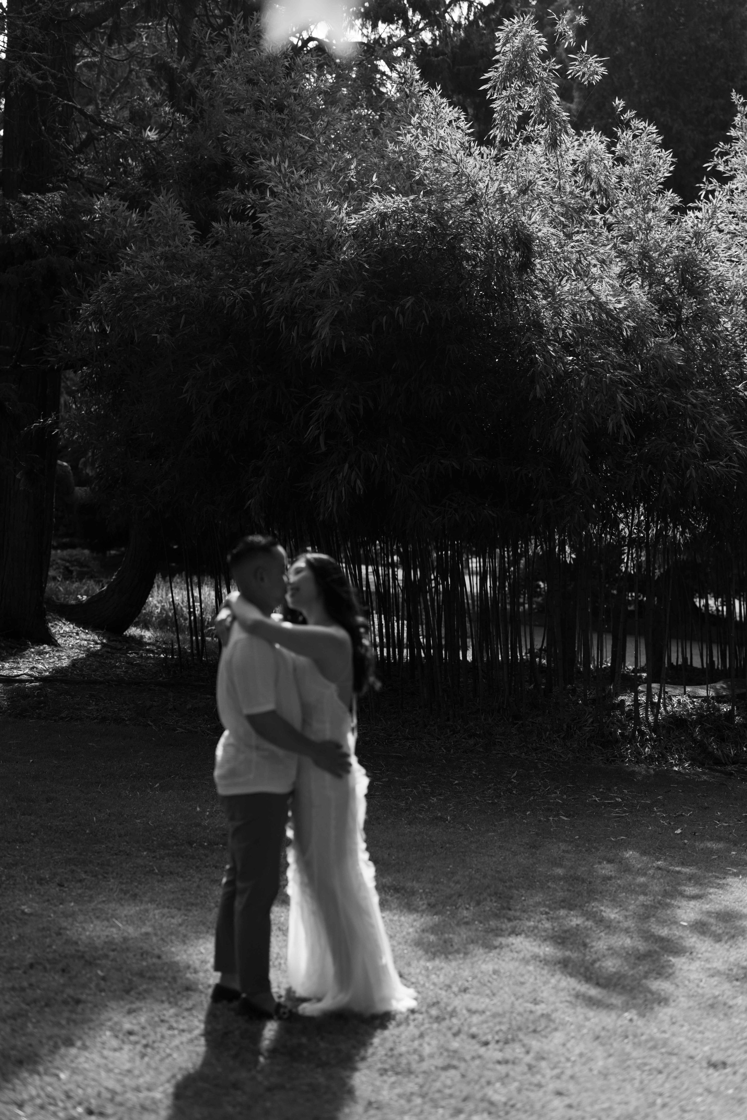Black and white photo of a couple embracing in a lush garden, dressed in wedding attire, sharing a romantic moment.