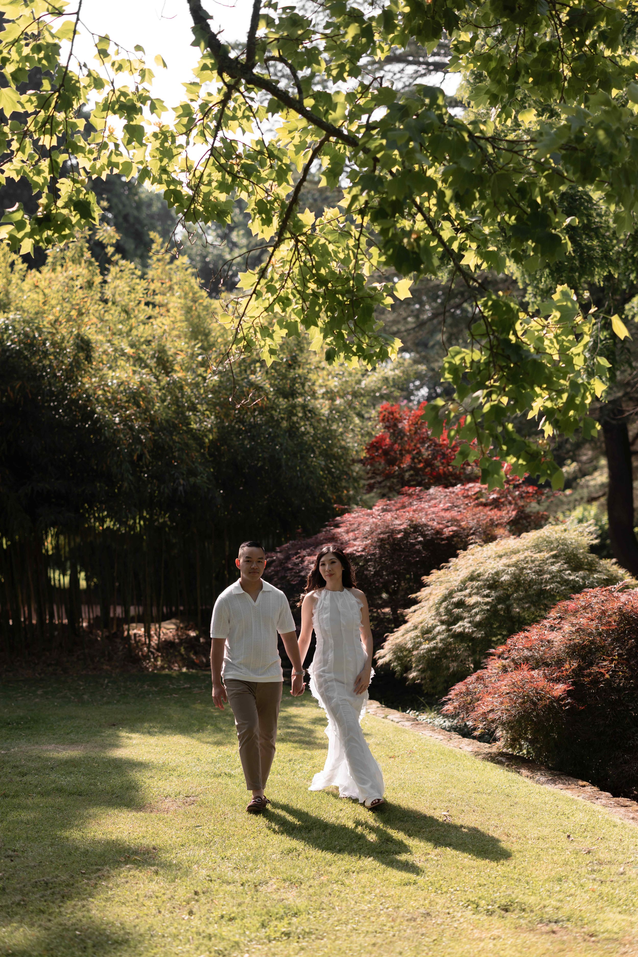 Couple in white attire holding hands while walking through a lush garden with colorful foliage on a sunny day.