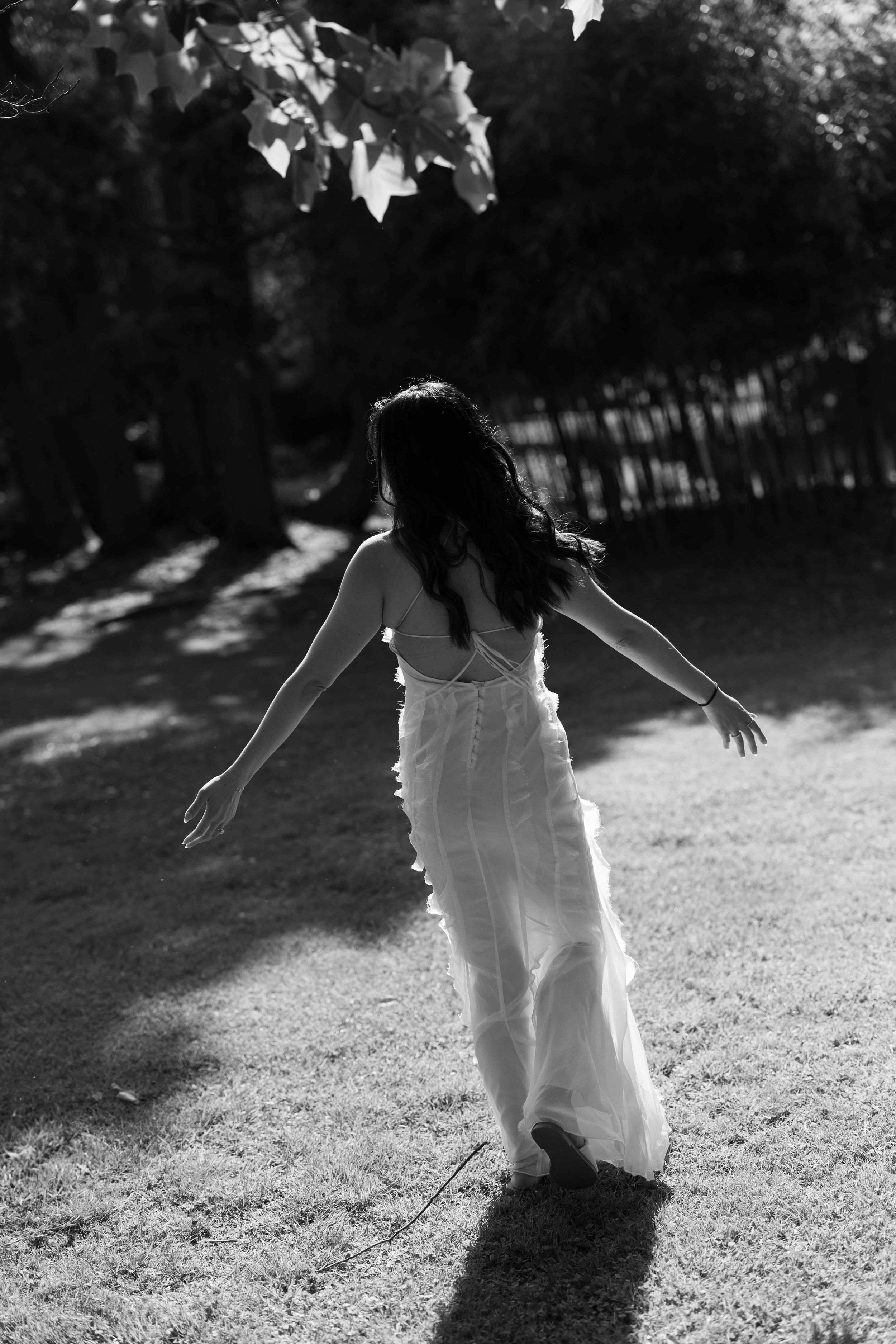 Black and white photo of a woman in a white dress walking through a sunlit park with arms outstretched.