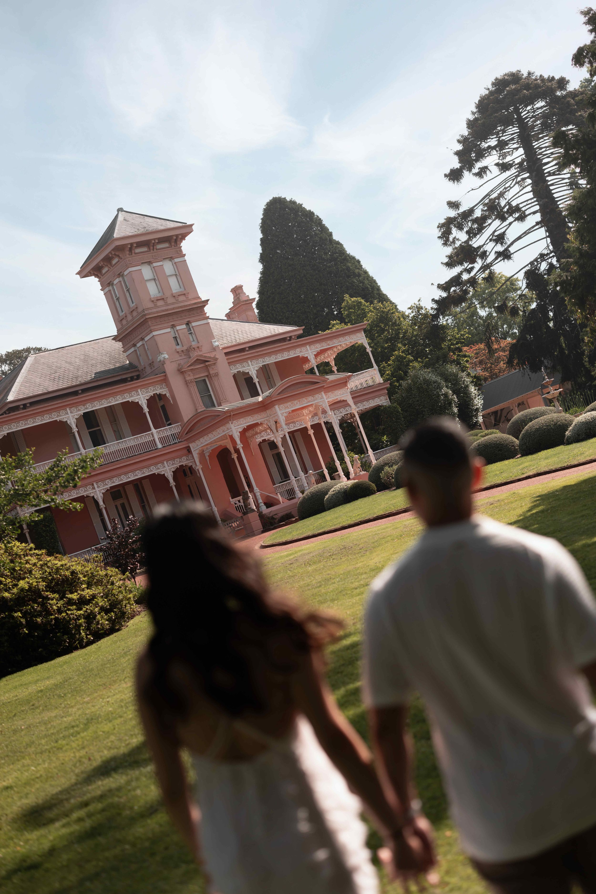 Couple holding hands walking toward a grand pink Victorian mansion surrounded by lush green gardens on a sunny day.