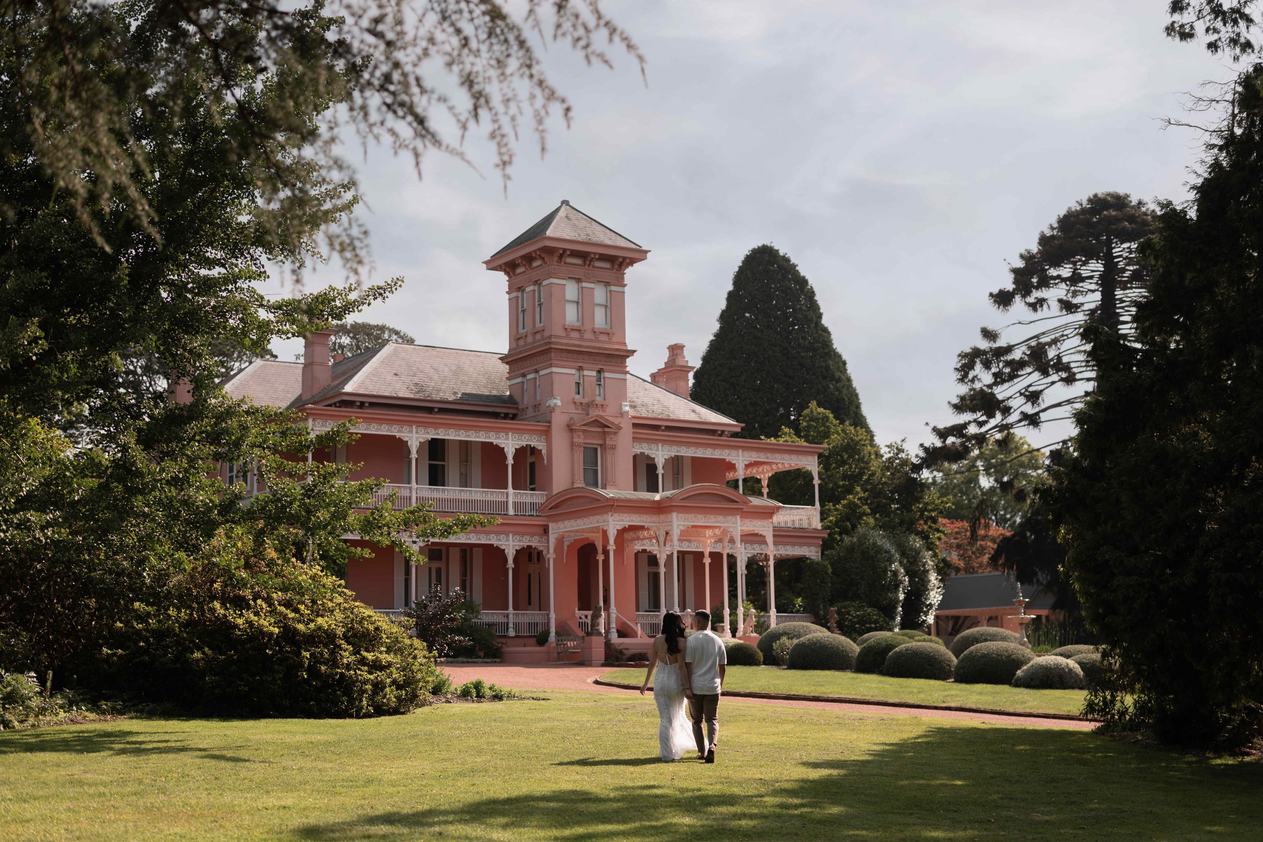Couple walking toward a grand pink Victorian mansion surrounded by lush green gardens and tall trees.