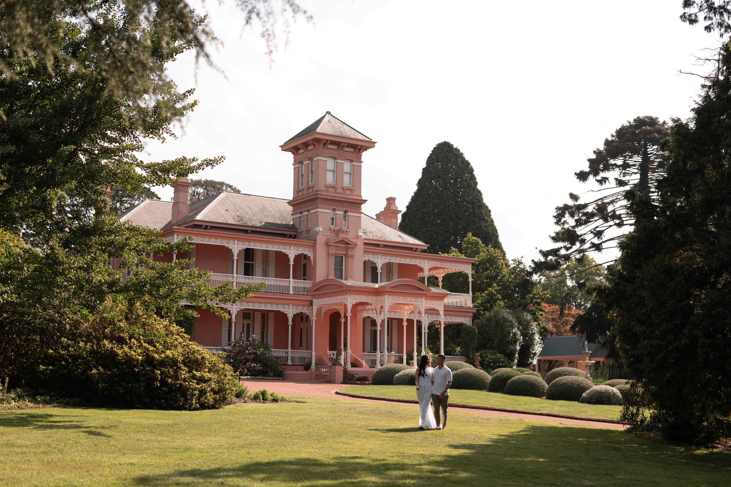 Couple walking toward a grand pink Victorian mansion with a tower, wrap-around porch, and lush green gardens.