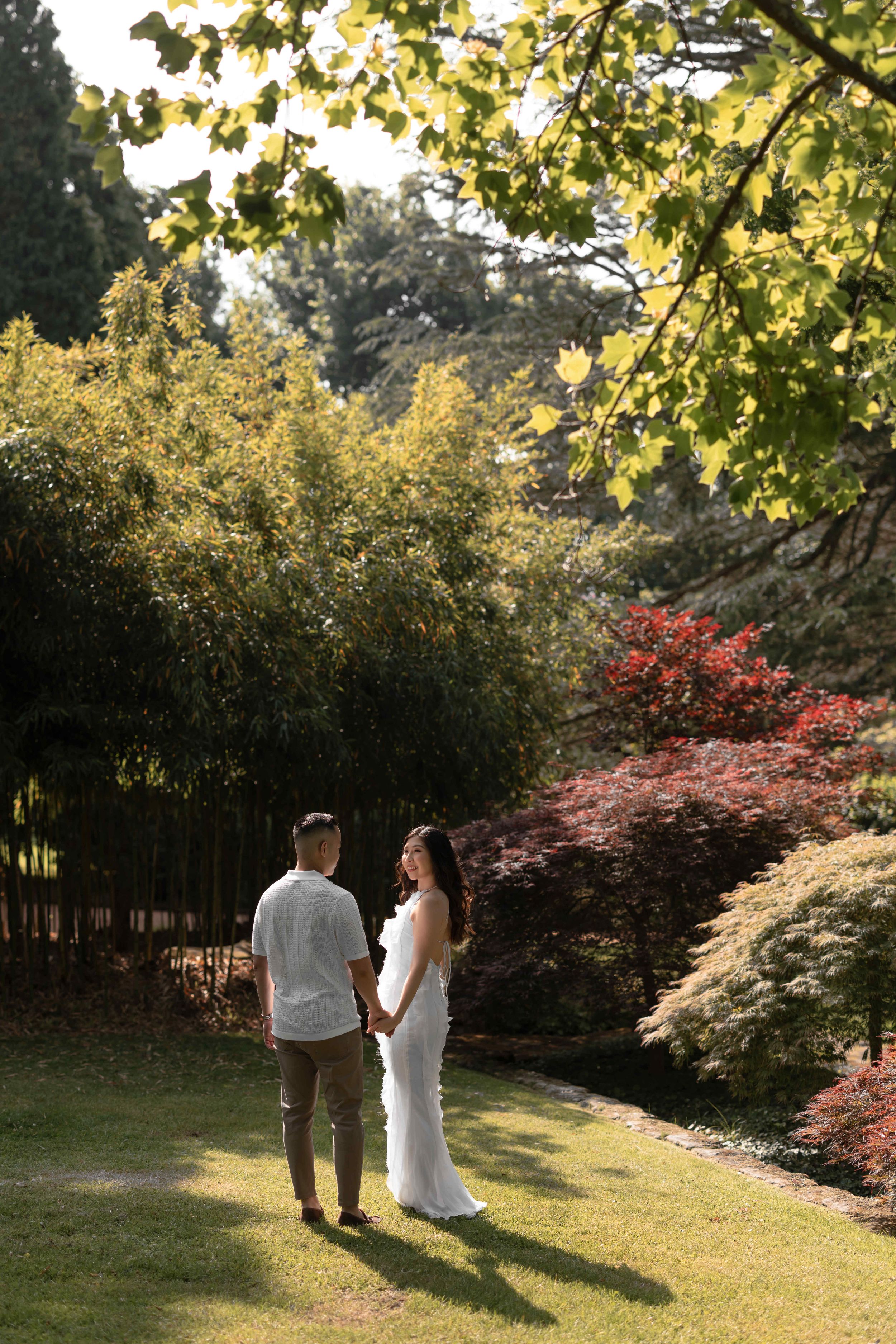 Couple holding hands in a lush garden, woman in white dress, surrounded by colorful foliage in warm sunlight.