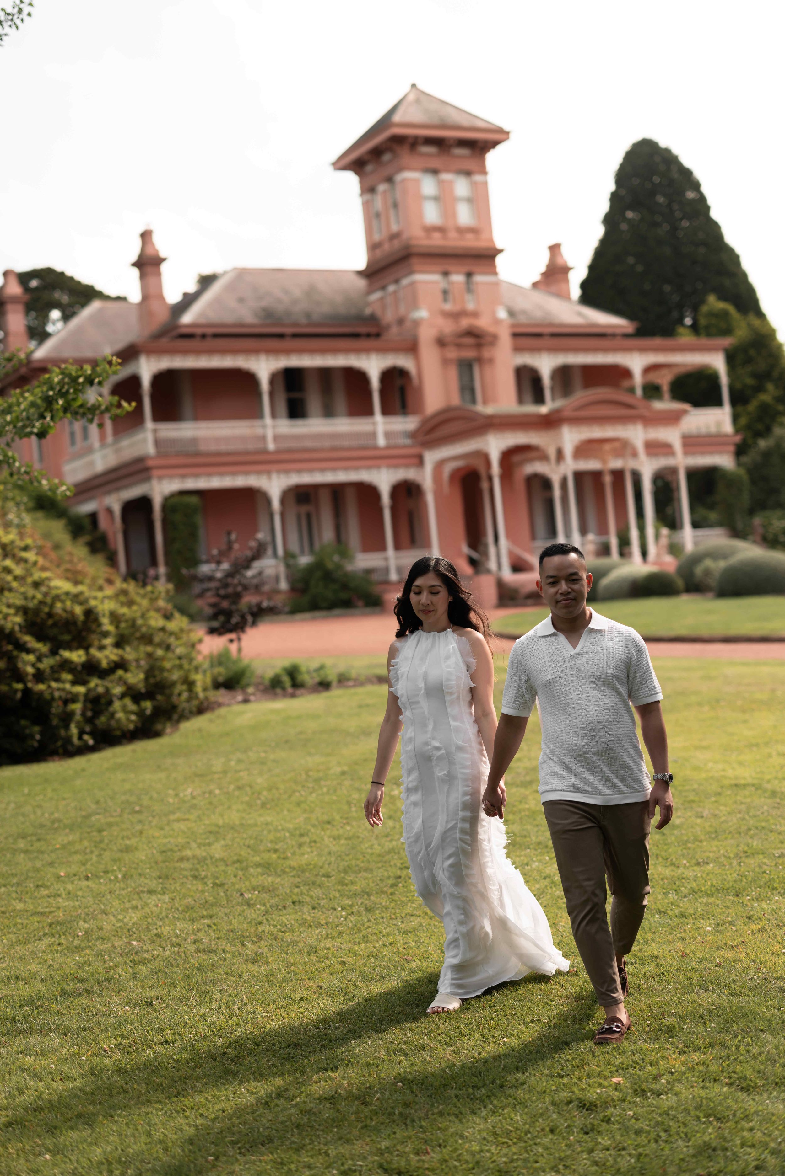 Couple holding hands walking on green lawn in front of a grand pink Victorian mansion with wrap-around veranda.
