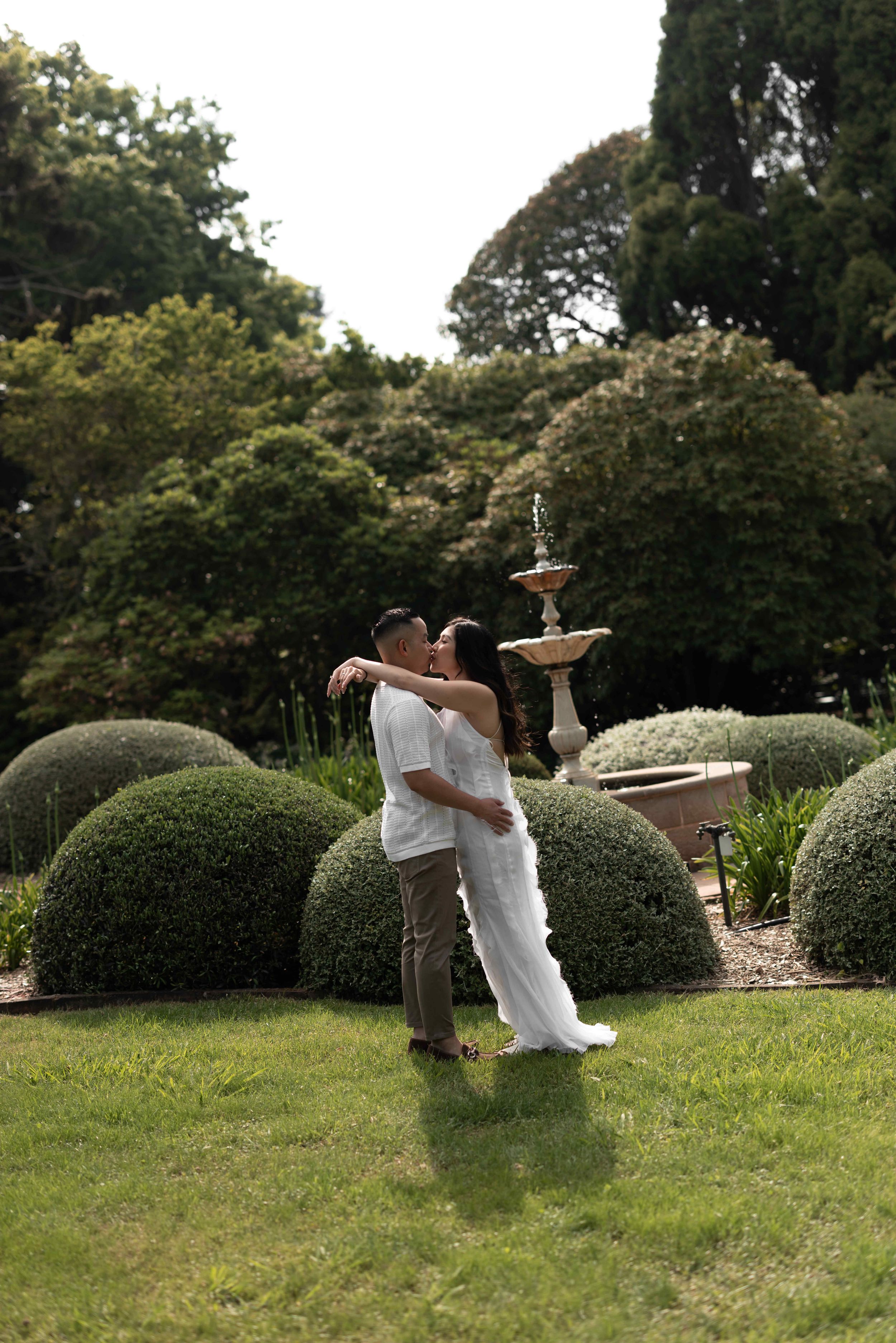 Couple sharing a romantic kiss in a lush garden with trimmed hedges and a stone fountain backdrop.