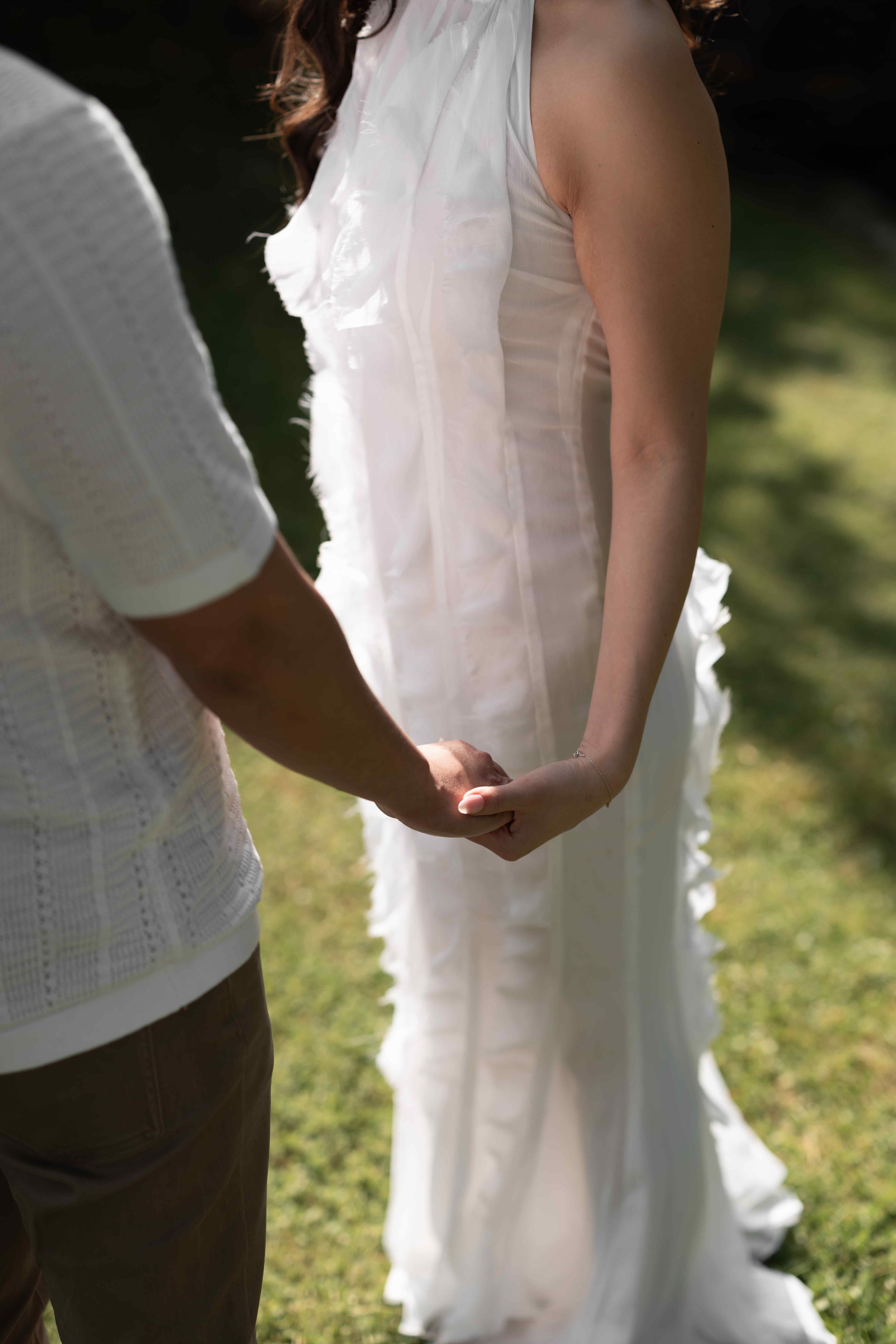 Bride in white wedding gown holding hands with groom outdoors on green grass in sunlight.