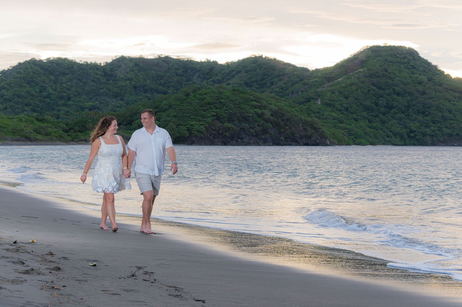 Couple walking hand in hand on a tropical beach at sunset with lush green hills in the background.