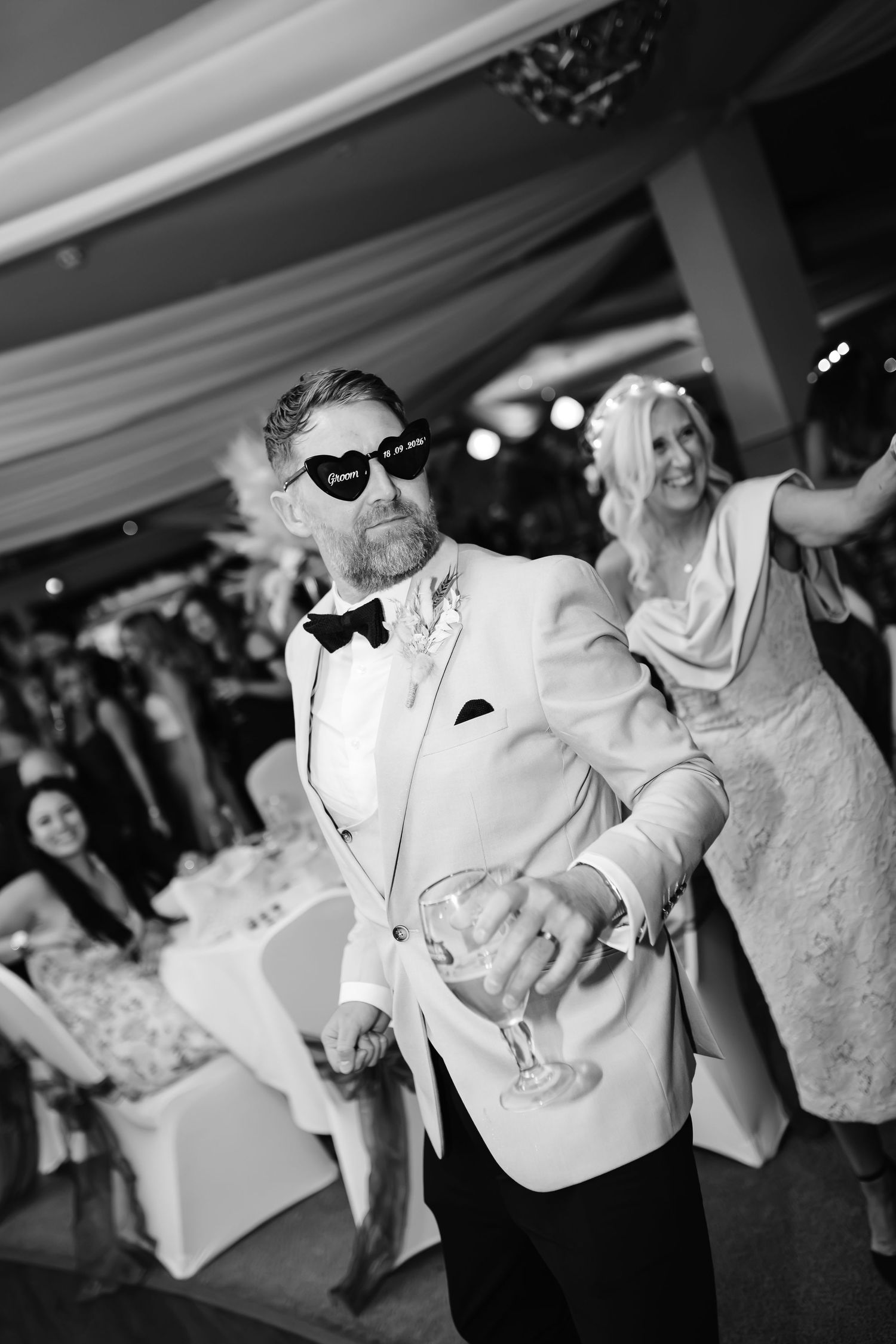 Bride in a strapless gown and groom in a blazer and bow tie dance playfully in sunglasses at their wedding reception.