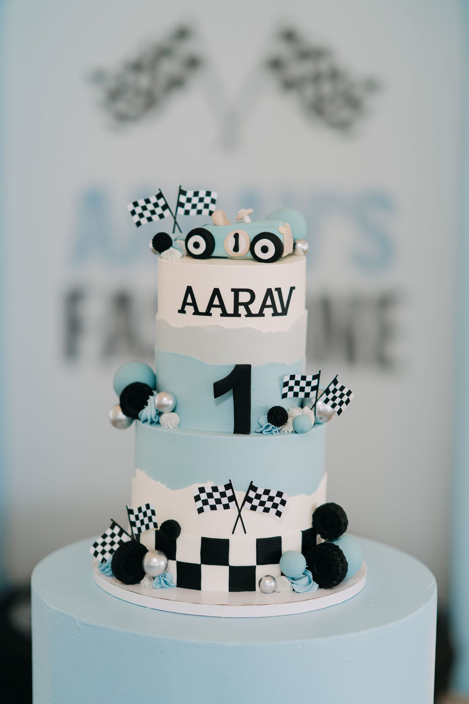 Light blue first birthday cake with Aarav name topper, race car, checkered flags, black and silver decorations, and a number one.