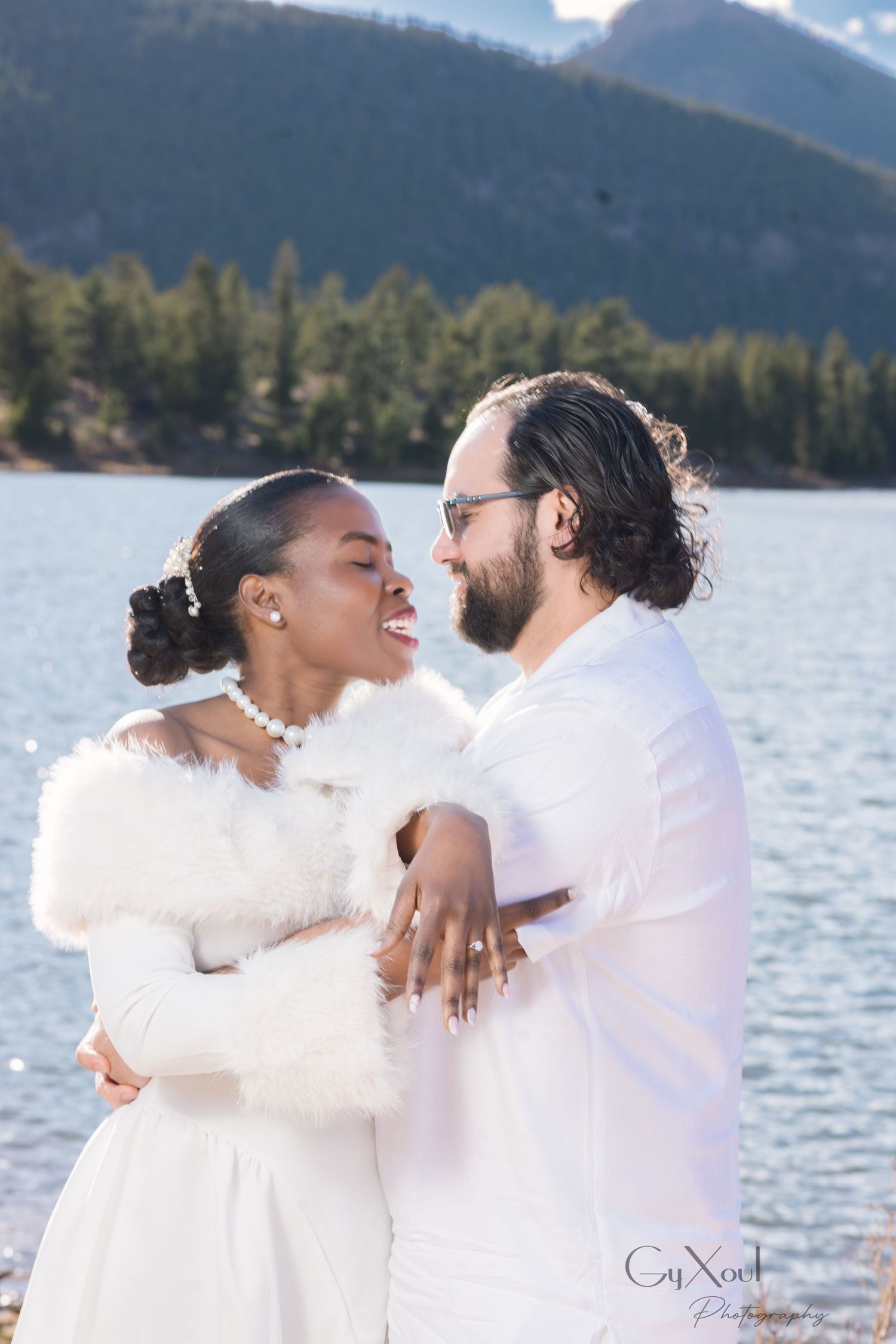 Smiling couple in white embracing beside a mountain lake, the bride wearing a faux fur wrap, with evergreen forest and mountains behind.