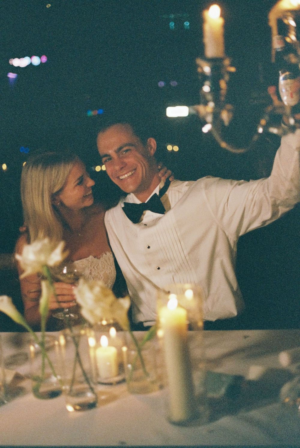 Smiling groom in a white dinner jacket and bow tie takes a selfie with the bride beside candles and white flowers at night.