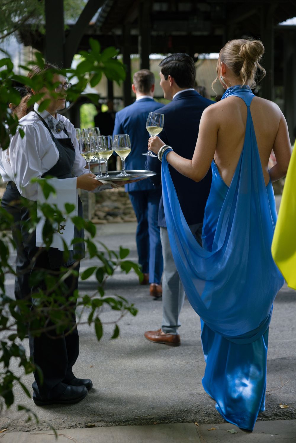 Woman in a flowing blue backless gown takes a glass of white wine from a waiter's tray at an outdoor garden reception.