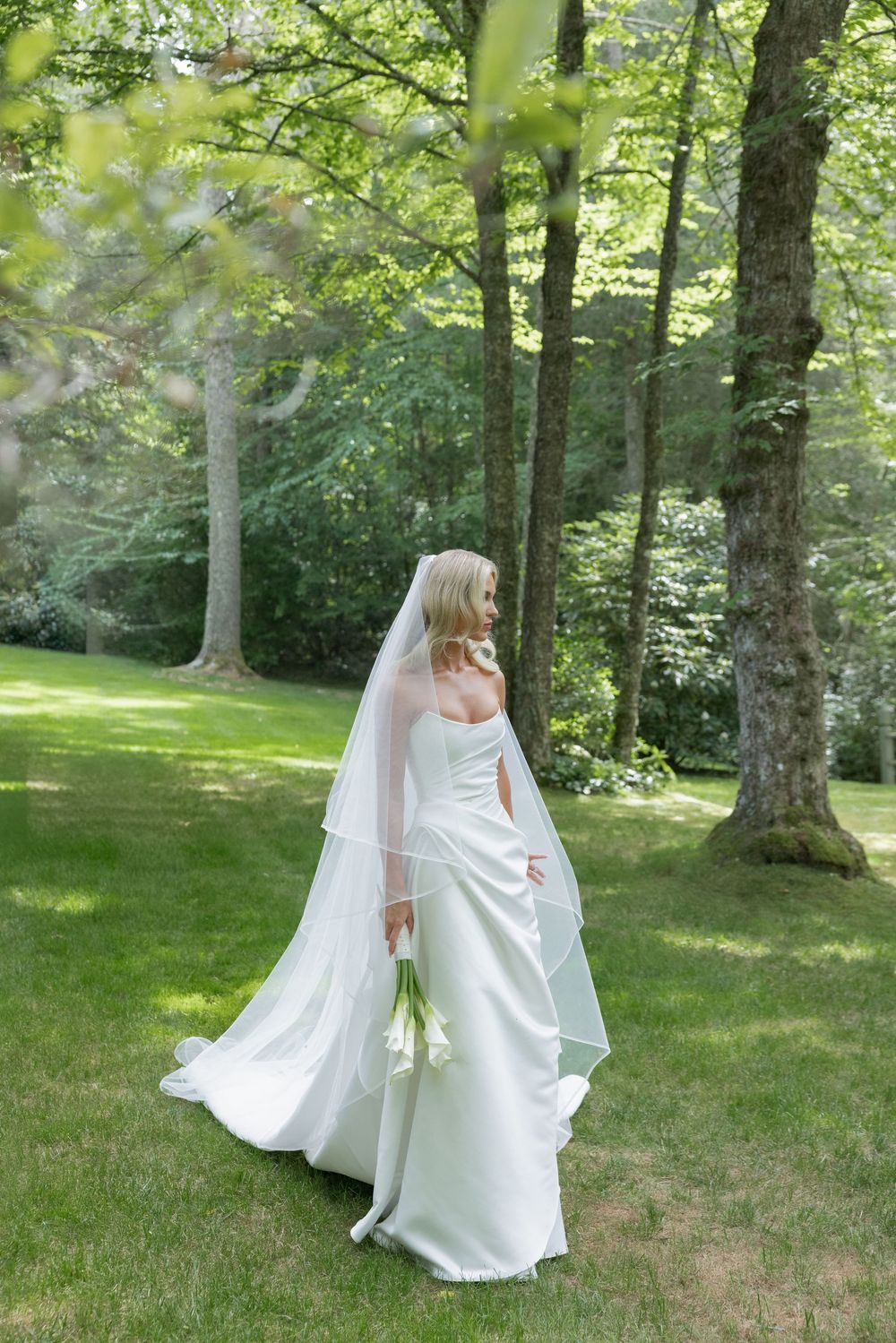 Bride in a satin gown and long veil holds white calla lilies while standing on a sunlit lawn among tall trees.