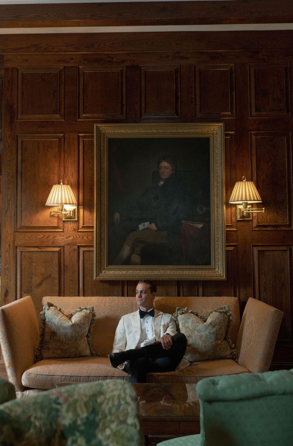 Man in a white dinner jacket and bow tie sits on a tan sofa beneath a framed portrait in a wood-paneled room with wall sconces.