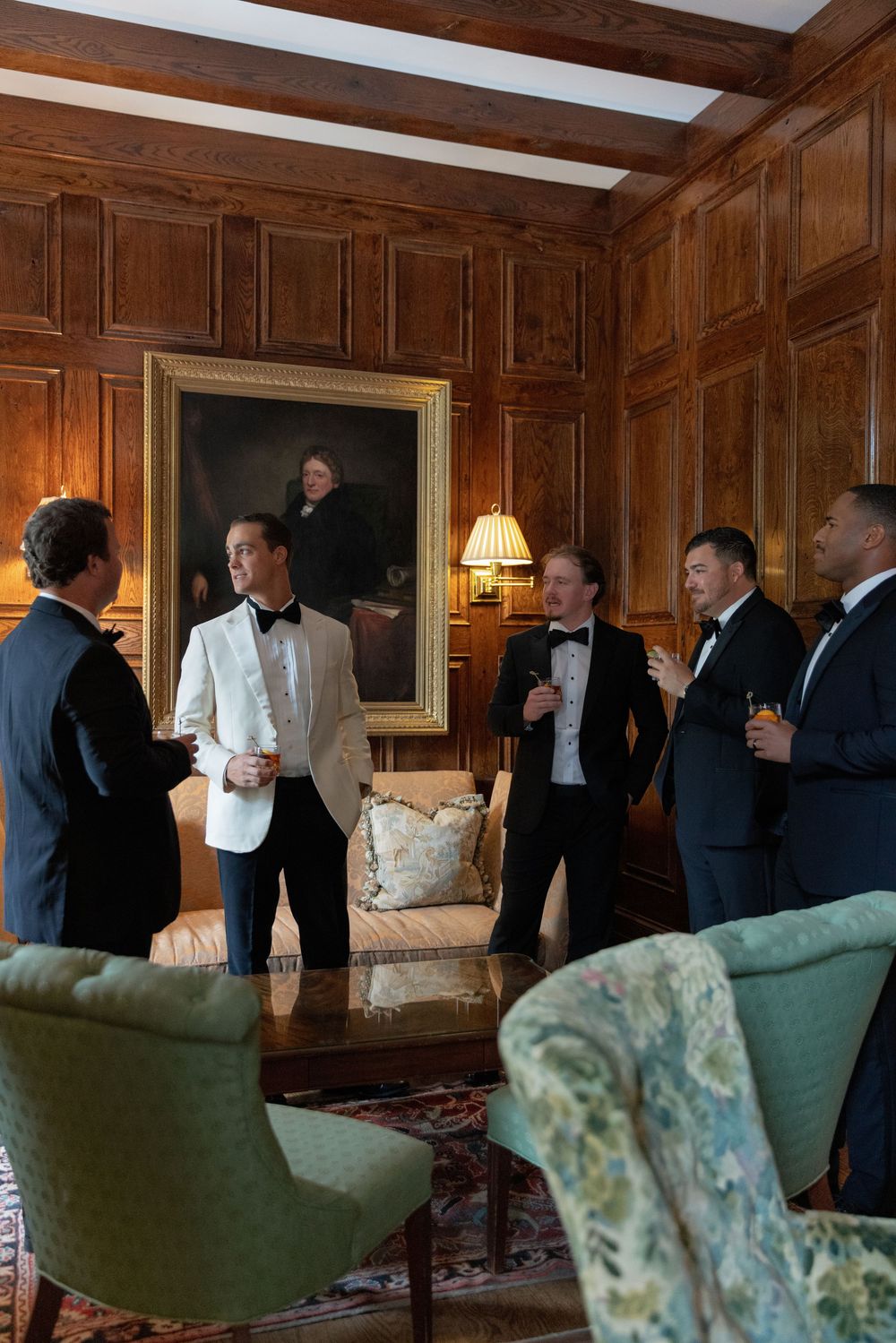 Groomsmen in tuxedos and a groom in a white dinner jacket chat with drinks in a wood-paneled room with a portrait.