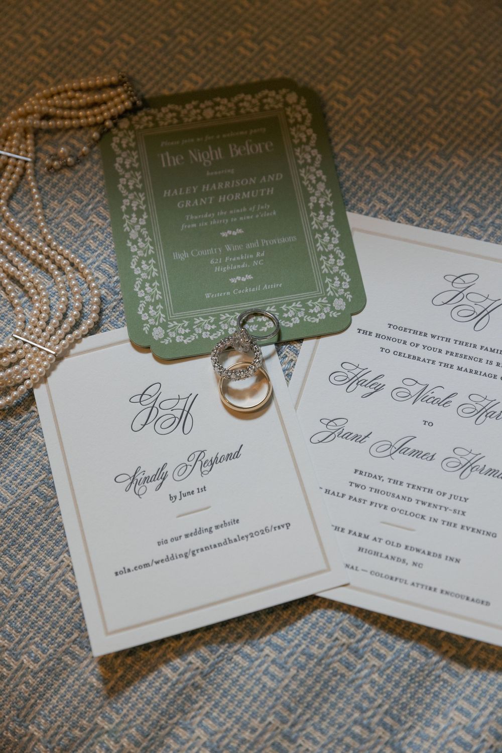 Wedding invitation suite with a green rehearsal dinner card, RSVP card, and engagement rings resting on a pearl necklace.