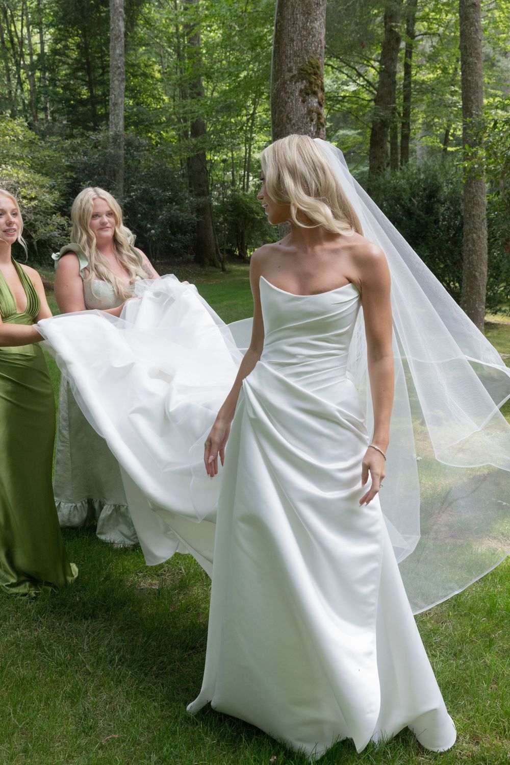Bride in a strapless satin wedding gown and long veil stands on grass as bridesmaids in green dresses lift her train in a sunlit forest.