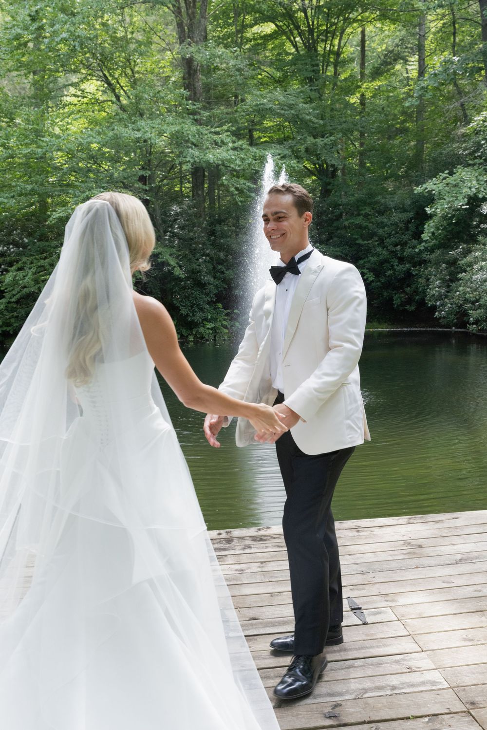 Groom in a white dinner jacket reaches for the bride's hand during a first look on a wooden dock by a lake and fountain.