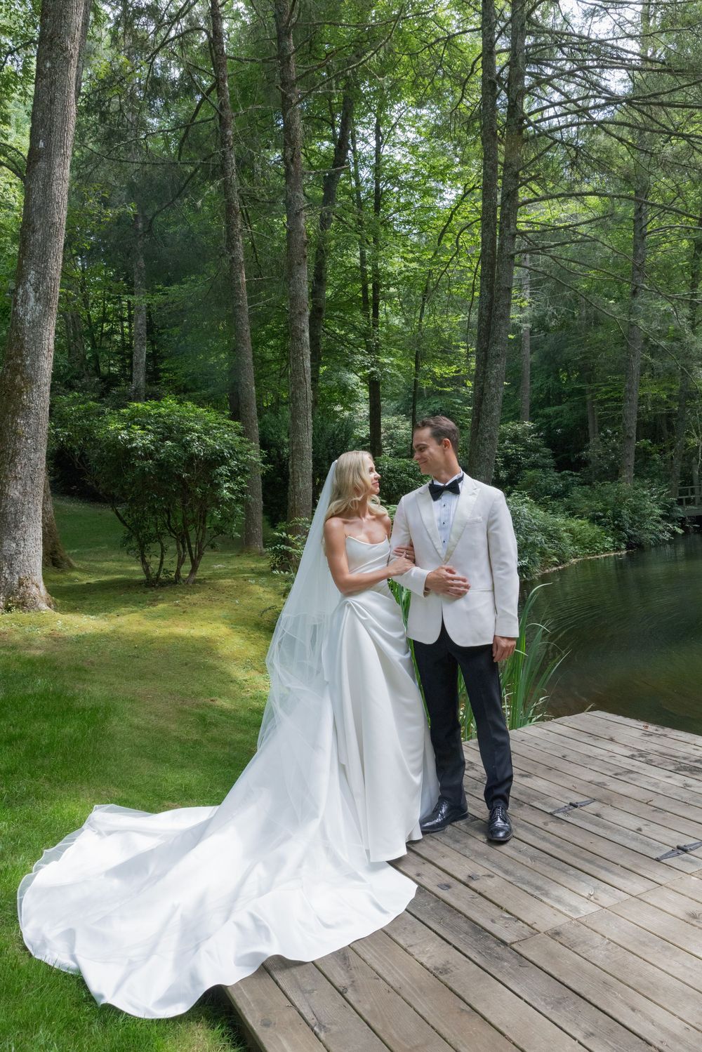 Bride in a flowing white gown and groom in a white jacket and bow tie embrace on a wooden dock beside a lake surrounded by lush green trees.