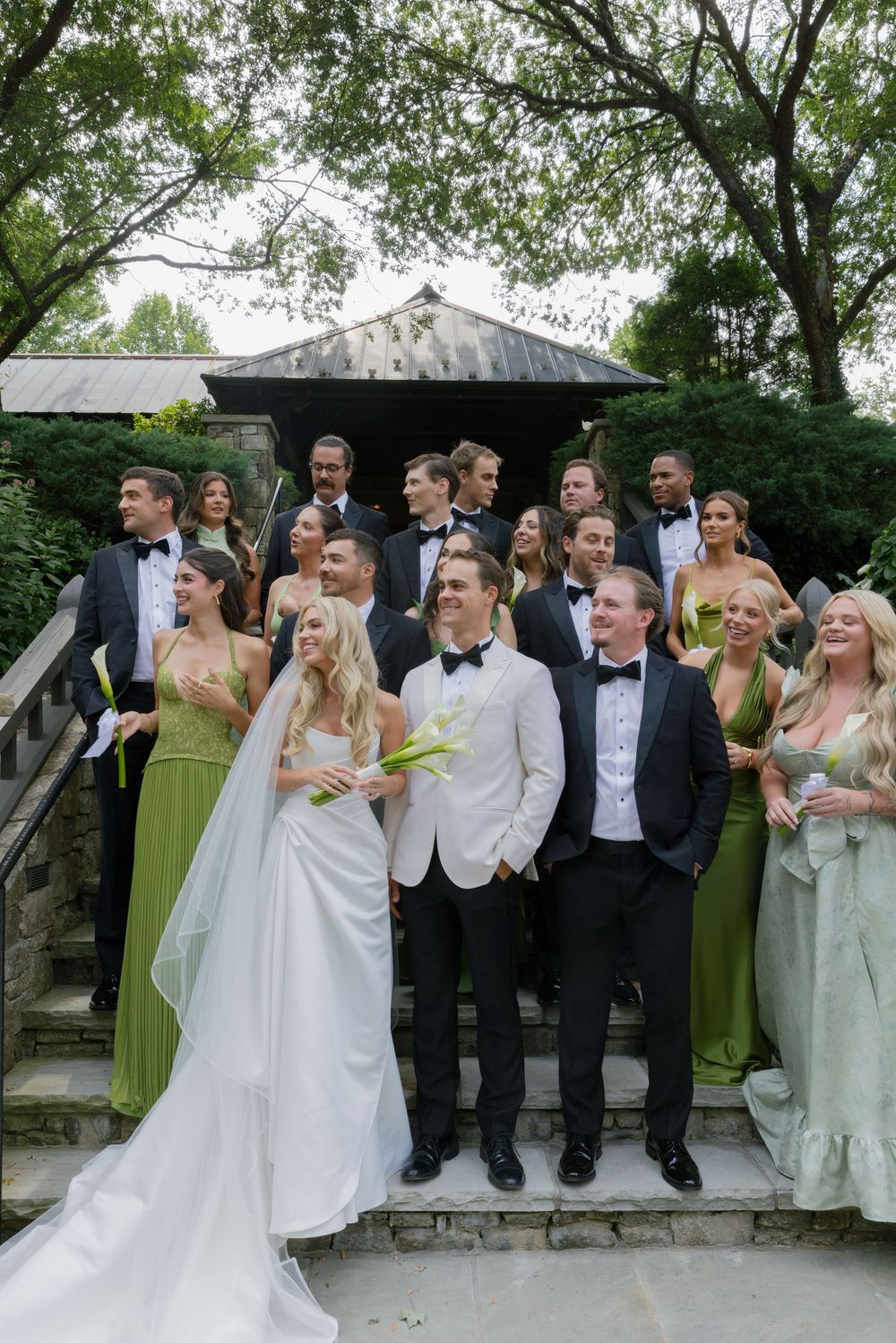 Bride and groom pose with bridesmaids and groomsmen in green dresses and tuxedos on stone steps surrounded by trees.