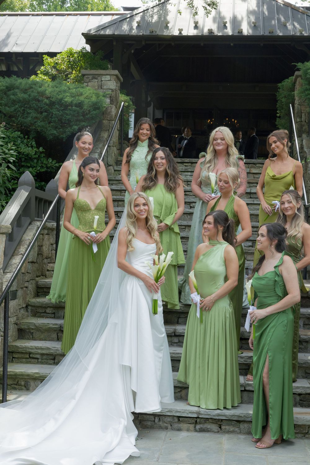 Bride in a white gown and veil poses with bridesmaids in green dresses on stone steps at an outdoor wedding venue.