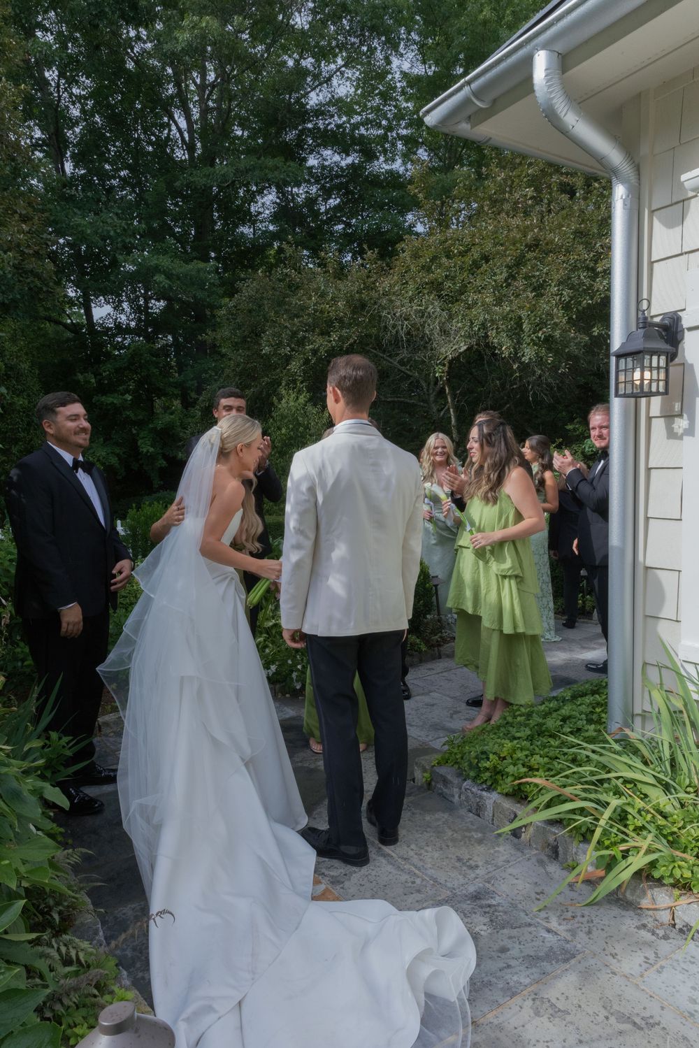Bride in a long white gown and groom in a white dinner jacket stand outdoors with wedding guests gathered beside a garden path.