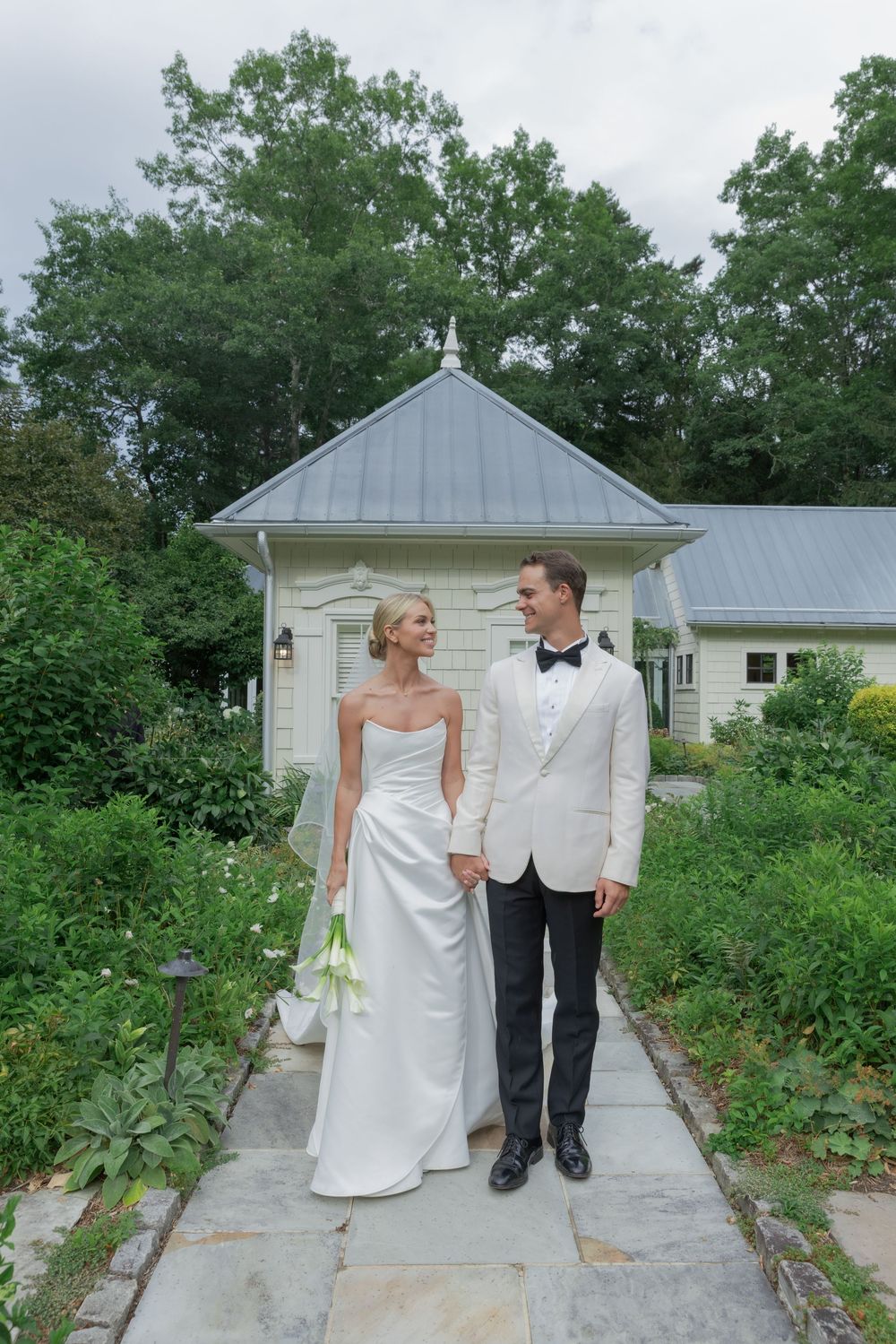 Bride and groom in a white dinner jacket and bow tie hold hands on a stone path near a garden cottage.