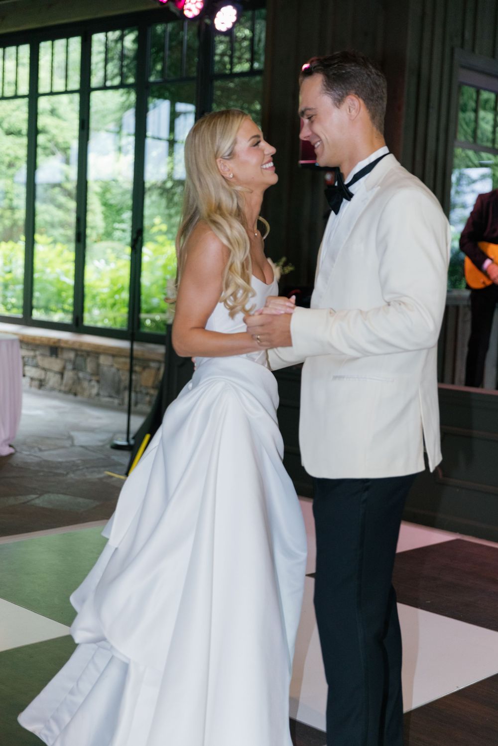 Smiling bride and groom share their first dance on a checkered floor in a rustic venue with large windows.