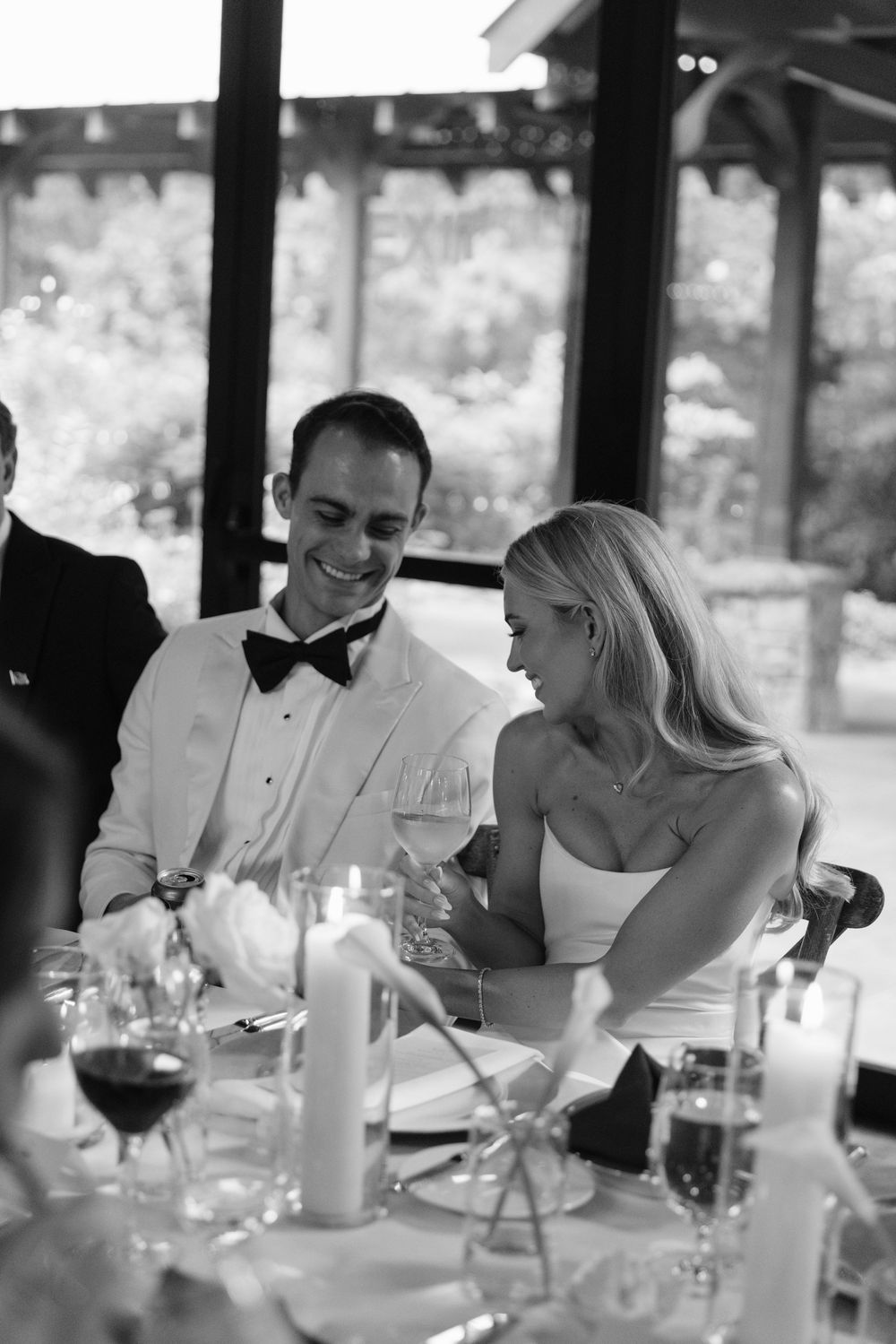 Black and white photo of the smiling bride and groom seated at their reception table with candles and wine glasses.