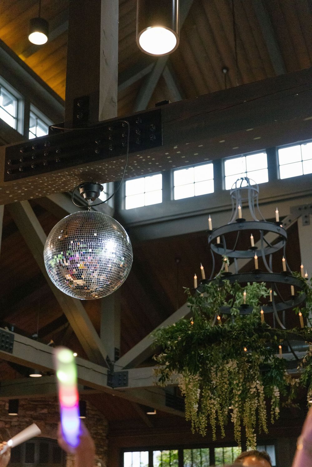 Disco mirror ball hangs beneath wooden beams beside a candlelit chandelier draped in greenery at a rustic barn wedding reception.