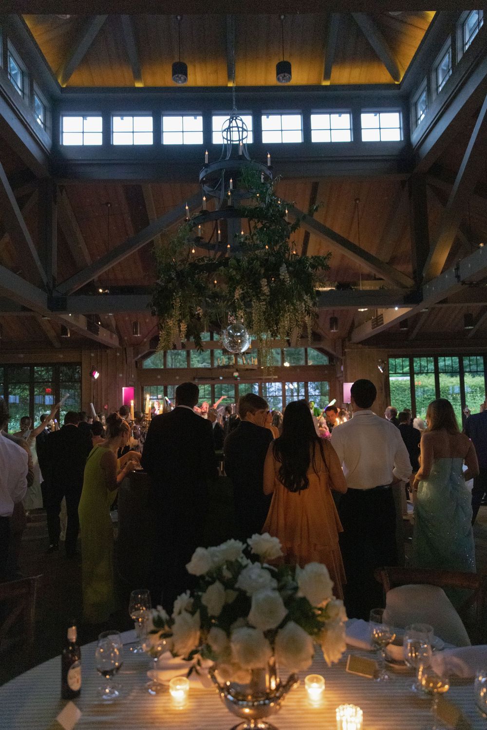 Wedding guests mingle in a rustic timber-framed venue with a hanging greenery chandelier and white floral centerpiece on a candlelit table.