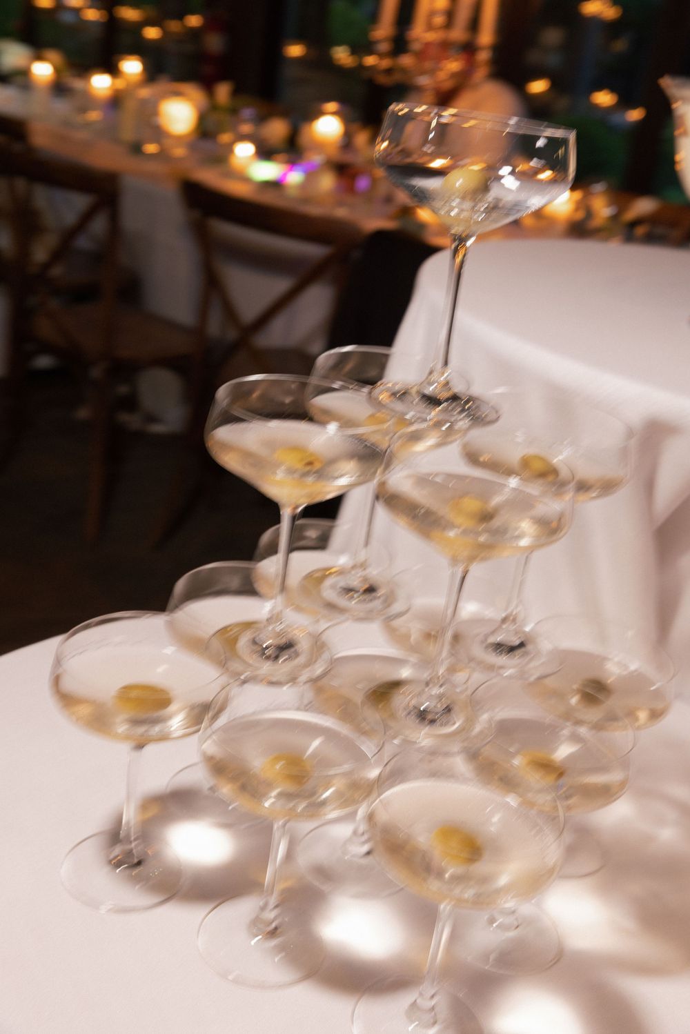 Tower of martini glasses garnished with olives sits on a white tablecloth with candlelit tables glowing in the background.