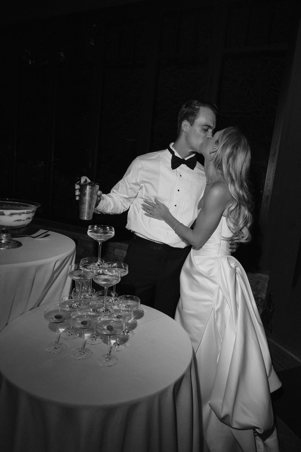 Black and white photo of a bride and groom kissing beside a tall champagne coupe tower on a draped table at their wedding reception.
