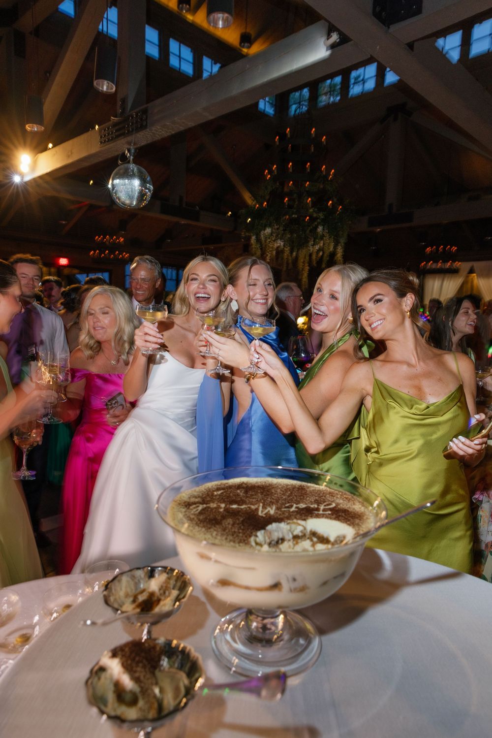 Bride celebrates with bridesmaids holding cocktails around a table with tiramisu desserts in a festive reception hall.