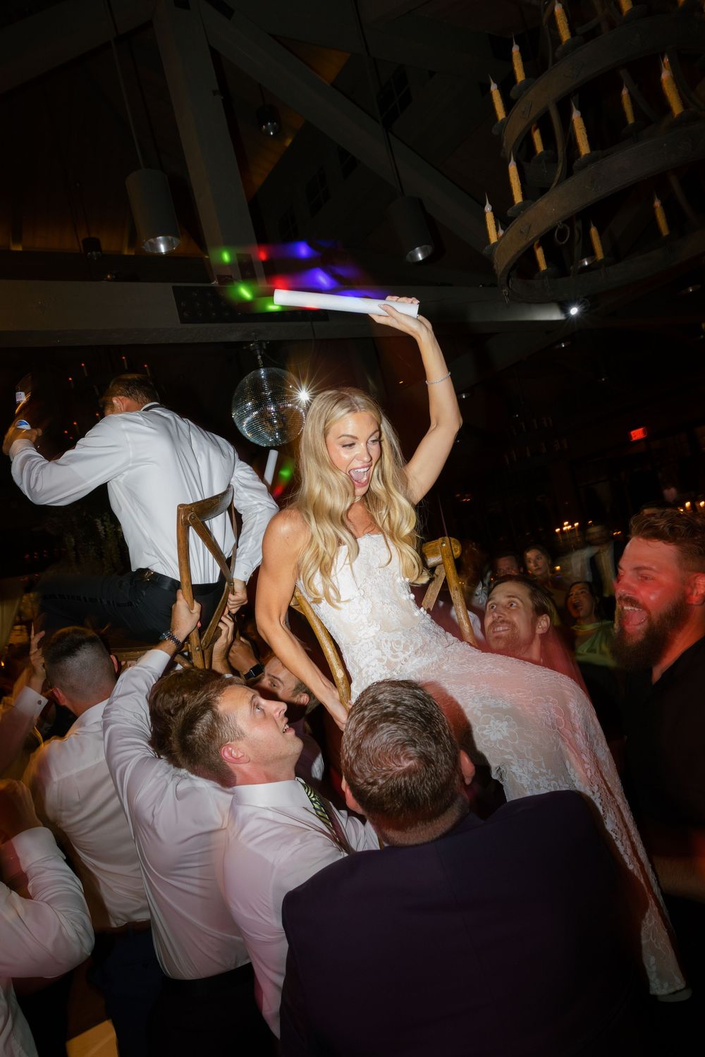 Guests lift the bride in her wedding gown on a chair during an energetic dance floor celebration at the reception.