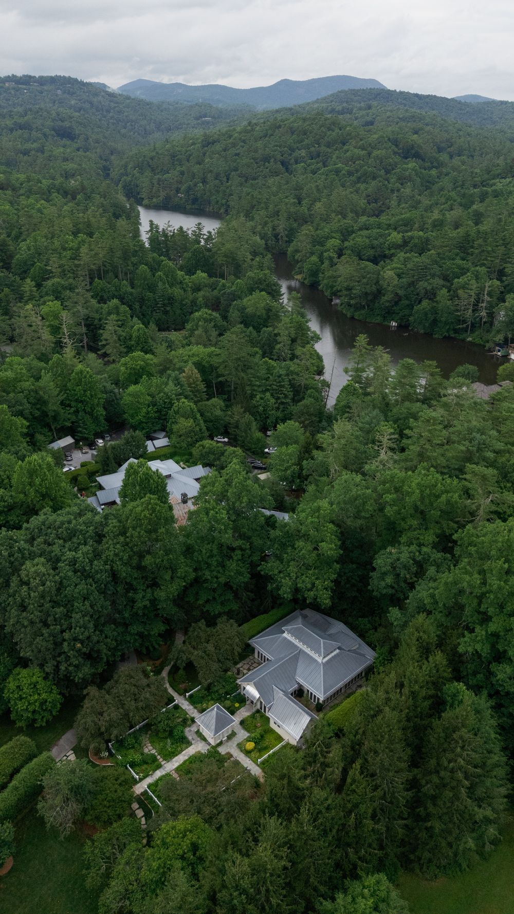 Aerial view of a gray-roofed mountain home surrounded by dense green forest, with a winding river and misty hills in the distance.