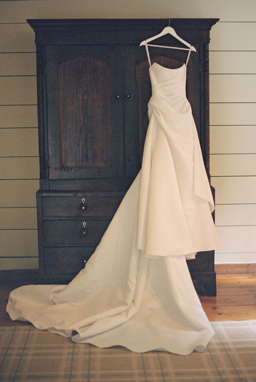 Ivory satin wedding gown with a ruched bodice and long flowing train hanging on a wooden hanger from a dark antique armoire.