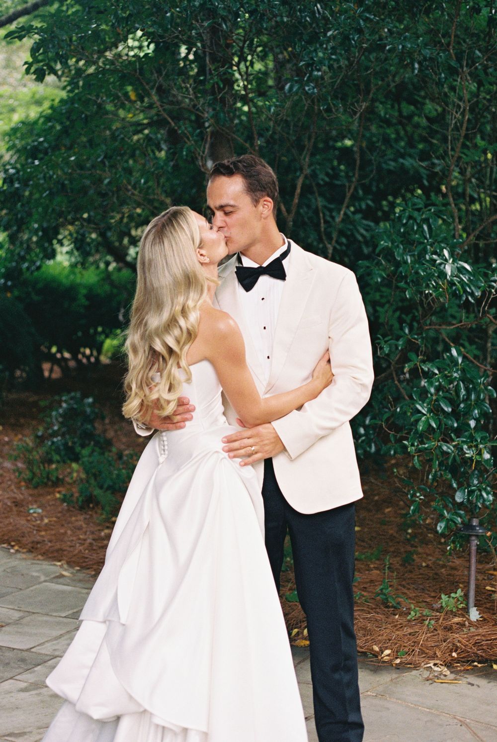 Bride in a flowing white gown and groom in a cream tuxedo jacket and bow tie share a kiss in a lush green garden.